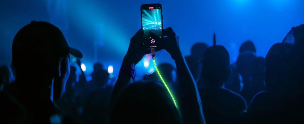Büyük bir kalabalığın önünde parlayan bir kabloya bağlı cep telefonunu yukarı kaldırıp etkinlikte anı yakalayan kişi